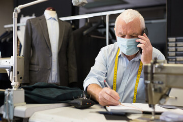 Mature tailor wearing protective mask talking on a mobile phone and taking notes in a notebook