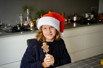 Teen girl in santa hat with chocolate cookie lollipop sitting at the table at home, enjoying Christmas holidays. Decoration with Christmas lights and ornaments.