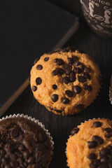 Delicious vanilla and chocolate muffins with chocolate chips next to a cup of coffee and a black cover book on a black background on a dark wooden table