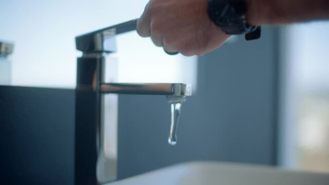A hand of a Caucasian male comes into frame as he opens the tap of a modern bathroom with the water starting to run.
