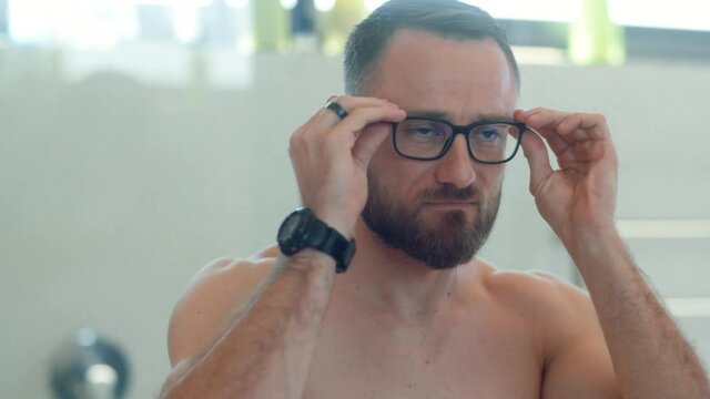 A bearded attractive Caucasian shirtless man wearing a towel around his waist whit a tattoo puts glasses on his face, checks himself out and proceeds to walk out of frame in a modern bathroom.