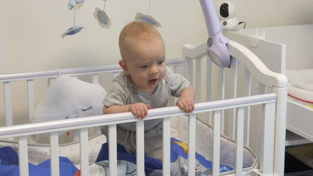 Happy cute adorable infant baby boy standing holding hands on crib playing in white rocking cradle at home. High quality 4k footage