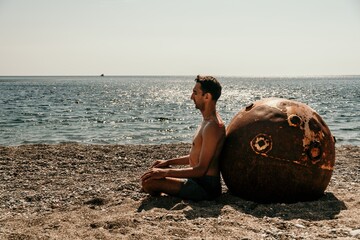 Man doing yoga and meditation outdoors near to old rusty floating marine mine on the beach with rocky shore and sea background. Healthy lifestyle, pollution, nature protection, war and peace concept. - Powered by Adobe