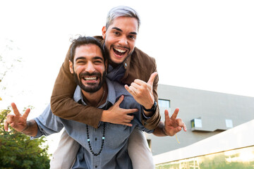 Playful smiling male gay couple looking at camera. Man piggyback ride with boyfriend. Copy space.