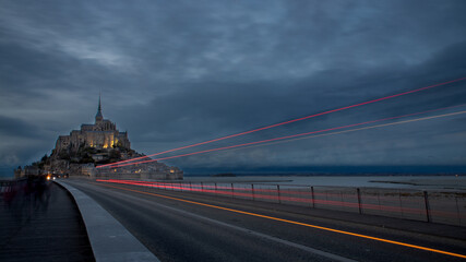 Fototapeta na wymiar Vue du Mont Saint Michel à l'heure bleue avec, en avant plan, les lignes de phares d'un bus