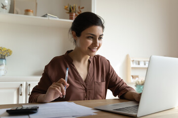 Indian woman sit at table look at laptop read online information, check e-bank statement, hold pen...
