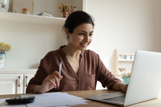 Indian woman sit at table look at laptop read online information, check e-bank statement, hold pen studying, e-learn use internet resources, working remotely, do paperwork, finances management concept