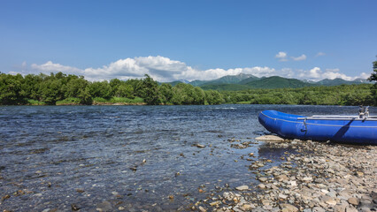 The river flows along a rocky bed. There is an inflatable boat for rafting on the shore. Lush green vegetation in the distance. A picturesque mountain range against the blue sky. Kamchatka