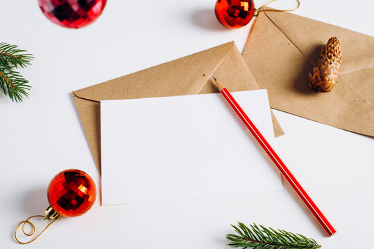 Merry Christmas festive composition layout. Close-up greeting card mock-up with empty space, craft envelope, pencil, red toy balls, pine cone and twigs on white table. Xmes, happy new year still life