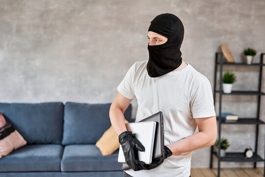 Thief with black balaclava stealing modern expensive laptop from apartment. Masked face. Man burglar stealing tv set from house