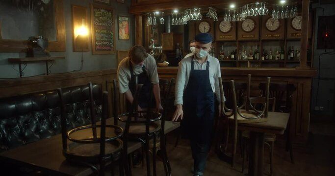 Barman and Staff in facial masks preparing restaurant for reopening after quarantine. Young waiters in aprons protective masks and gloves arranging furniture in cafe before open. The concept of