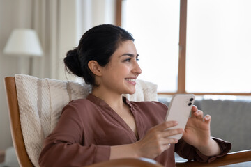 Dreamy beautiful Indian woman looks aside rest on cozy armchair with cellphone in hands daydreams enjoy carefree weekend alone with modern technology at cozy living room. Lifestyle, connection concept