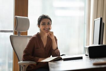 Smiling attractive Indian businesswoman sitting at desk in modern home office pose for camera feel...