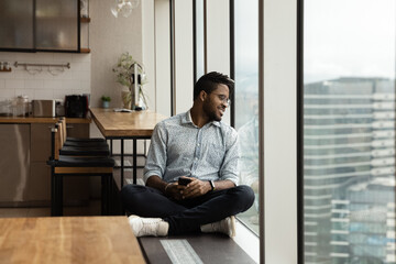 Relaxed carefree millennial African American latin hipster man sitting crossed legs on windowsill in modern office with smartphone in hands, looking outside admiring cityscape view in modern office.