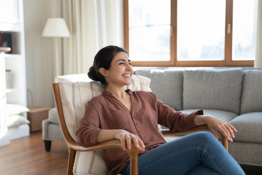 Happy Indian ethnicity woman relaxing on comfy wooden armchair spend time in cozy living room, smile enjoy carefree weekend and comfort furniture, breath fresh conditioned air inside modern apartment