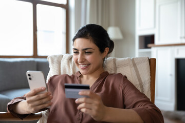 Fototapeta na wymiar Happy Indian woman sit on armchair holds debit card and smart phone buying on internet. Purchasing goods, retail services online, remote comfort usage of modern technology, secure e-bank app concept