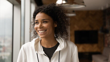 Happy attractive curly young African American woman in casual wear looking in distance, daydreaming, planning future or visualizing future standing in modern home office, inspiration concept.
