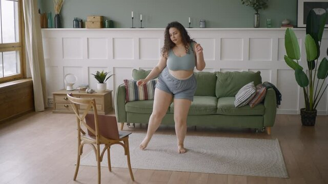 Curly loose haired plus size young woman in top and shorts watches online video on laptop and learns to dance on floor mat in spacious light room