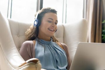 Happy young woman in wireless headphones looking at computer screen, watching funny movies or enjoying pleasant video call conversation, sitting in comfortable armchair, distant communication concept.