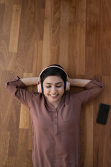 Vertical above view serene Indian woman lying indoor on laminate floor listen music through...