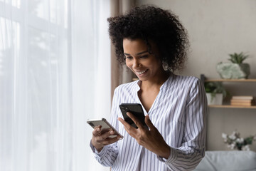 Smiling African American woman using two phones, standing at home, happy attractive young female...