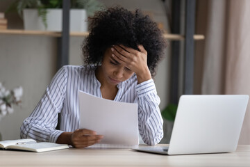 Unhappy African American woman reading bad news in letter, sitting at desk with laptop, upset...