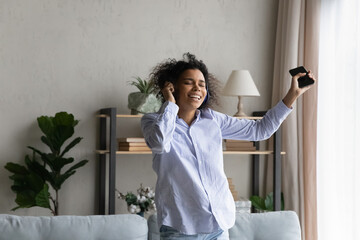 Excited African American woman in wireless headphones dancing at home, holding smartphone, listening to music relaxing in living room, happy young female in earphones enjoying leisure time weekend