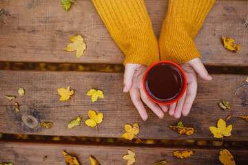 cup of hot tea in female hands holding it on wooden table autumn background with leaves. Warm drink concept.