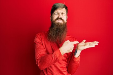 Redhead man with long beard pointing open palm with finger puffing cheeks with funny face. mouth...