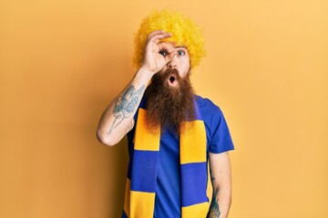 Redhead man with long beard football hooligan cheering game wearing funny wig doing ok gesture shocked with surprised face, eye looking through fingers. unbelieving expression.