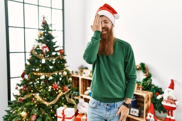 Redhead man with long beard wearing christmas hat by christmas tree covering one eye with hand, confident smile on face and surprise emotion.