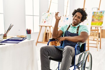 Young african american artist man sitting on wheelchair at art studio with a big smile on face, pointing with hand and finger to the side looking at the camera.