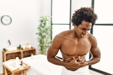 Young african american man shirtless wearing towel standing at beauty center with hand on stomach because nausea, painful disease feeling unwell. ache concept.
