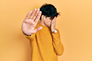 Handsome hipster young man wearing casual yellow sweater covering eyes with hands and doing stop gesture with sad and fear expression. embarrassed and negative concept.