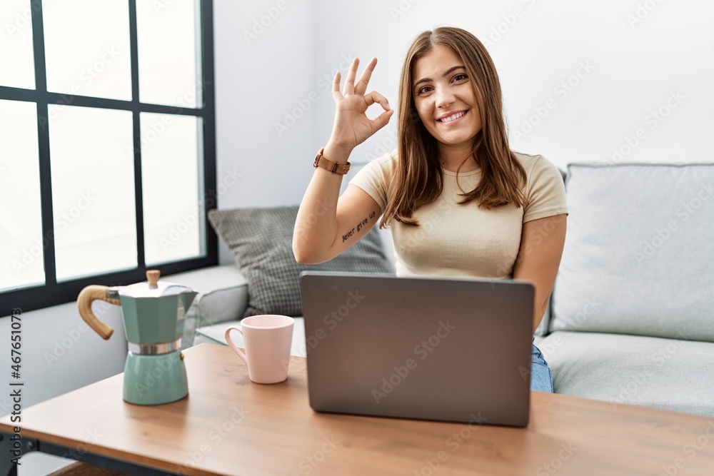 Canvas Prints Young brunette woman using laptop at home drinking a cup of coffee smiling positive doing ok sign with hand and fingers. successful expression.