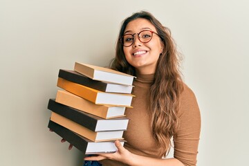Young hispanic girl holding a pile of books smiling with a happy and cool smile on face. showing...