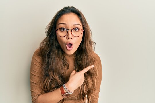 Young hispanic girl wearing casual clothes and glasses surprised pointing with finger to the side, open mouth amazed expression.