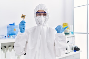 Young blonde woman holding cpu computer processor at laboratory pointing thumb up to the side smiling happy with open mouth