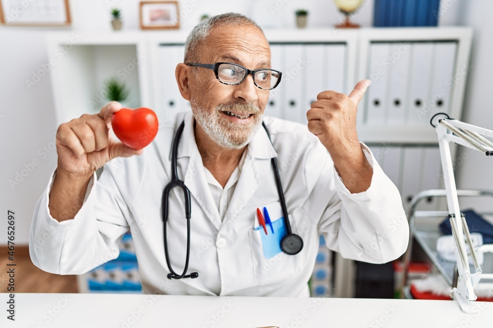 Wall mural Mature doctor man holding red heart at the clinic pointing thumb up to the side smiling happy with open mouth