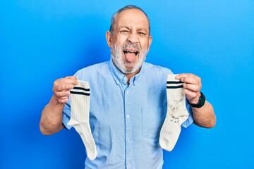 Handsome senior man with beard holding clean andy dirty socks sticking tongue out happy with funny...