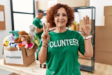 Middle age woman wearing volunteer t shirt at donations stand showing and pointing up with fingers...