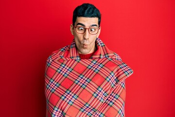 Fototapeta premium Handsome hispanic man wrapped in a red warm red blanket making fish face with mouth and squinting eyes, crazy and comical.