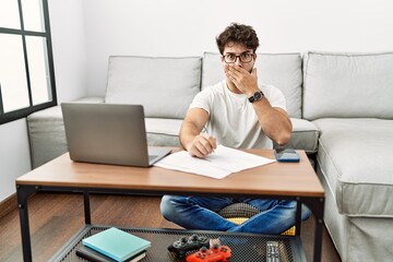 Hispanic man doing papers at home shocked covering mouth with hands for mistake. secret concept.