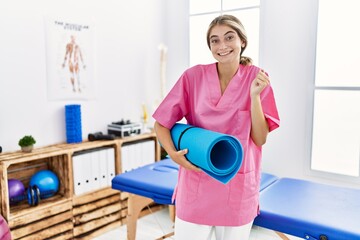 Young physiotherapist woman working at pain recovery clinic holding yoga mat screaming proud, celebrating victory and success very excited with raised arm