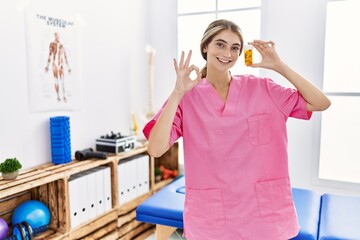 Young physiotherapist woman working at pain recovery clinic holding pills doing ok sign with fingers, smiling friendly gesturing excellent symbol