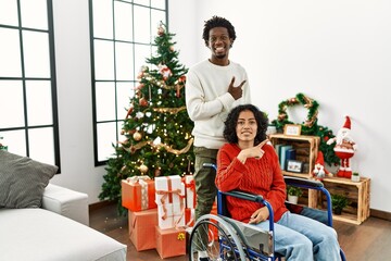 Young interracial couple with woman sitting on wheelchair by christmas tree cheerful with a smile of face pointing with hand and finger up to the side with happy and natural expression on face