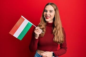 Young blonde woman holding hungary flag looking positive and happy standing and smiling with a confident smile showing teeth