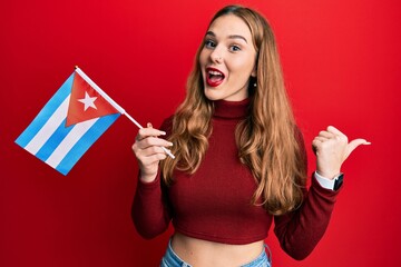 Young blonde woman holding cuba flag pointing thumb up to the side smiling happy with open mouth
