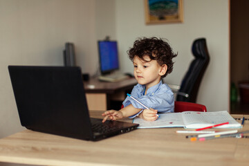 a happy preschool child is trained online on a laptop and does homework in a notebook. A cute, joyful curly boy looks at the monitor screen in a room of his house