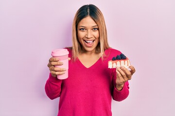 Beautiful hispanic woman holding takeaway cup of coffee and cheesecake smiling and laughing hard out loud because funny crazy joke.